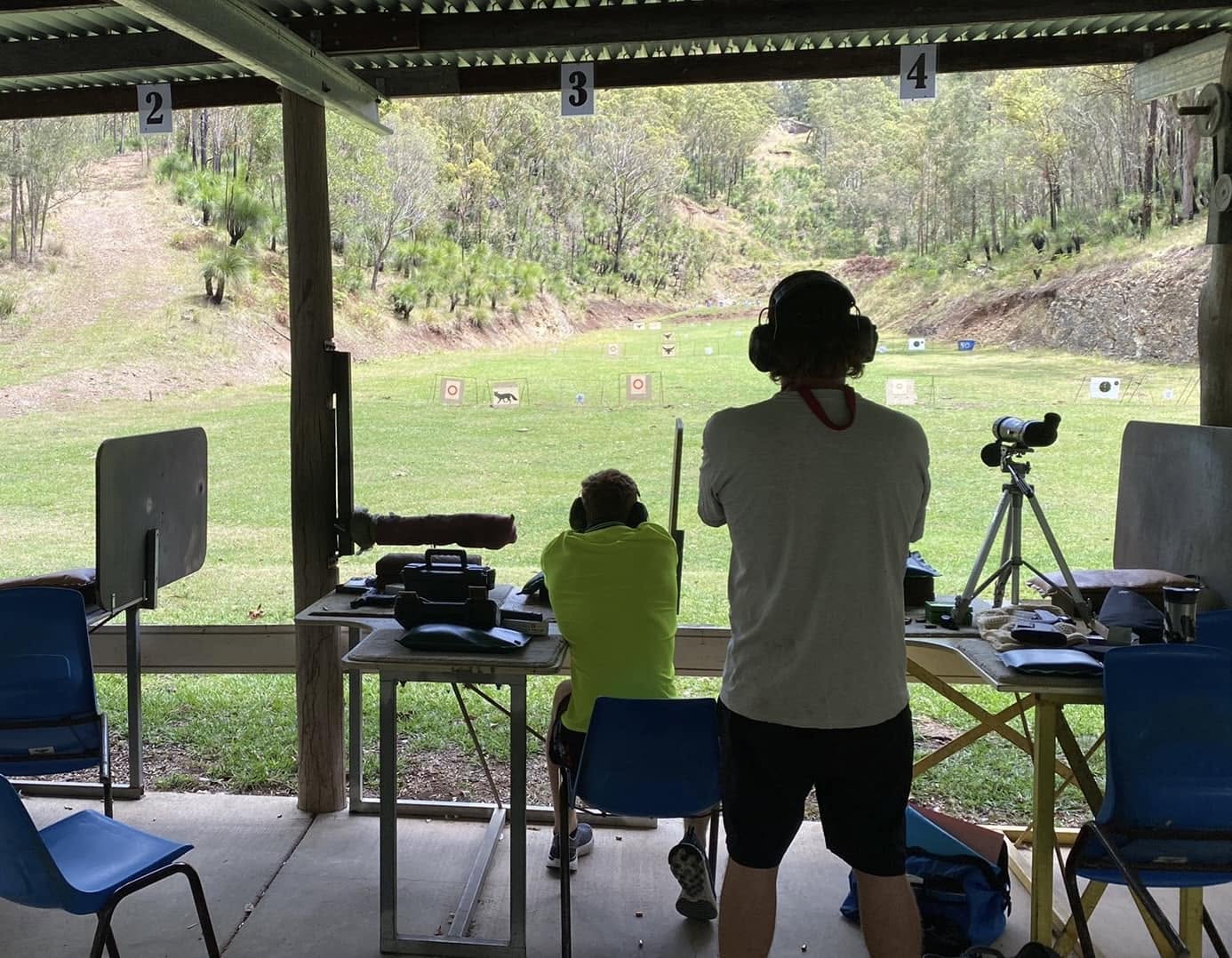 Shooters on the covered firing line at numbered benches, firing toward targets across the grassed range